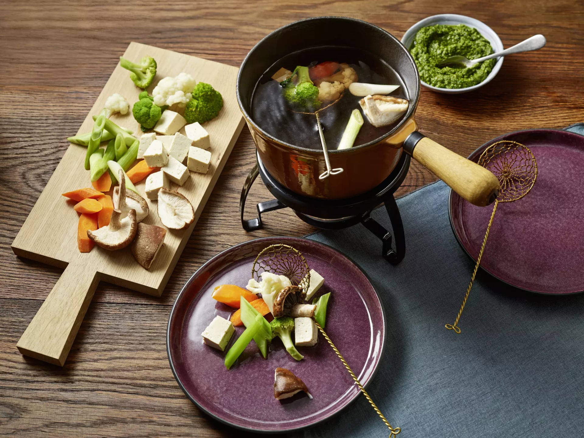 Fonduetopf mit Gemüse, daneben aufgeschnitten Pilze, Tofu, Brokkoli, Möhren Schale mit Pesto und zwei Teller.