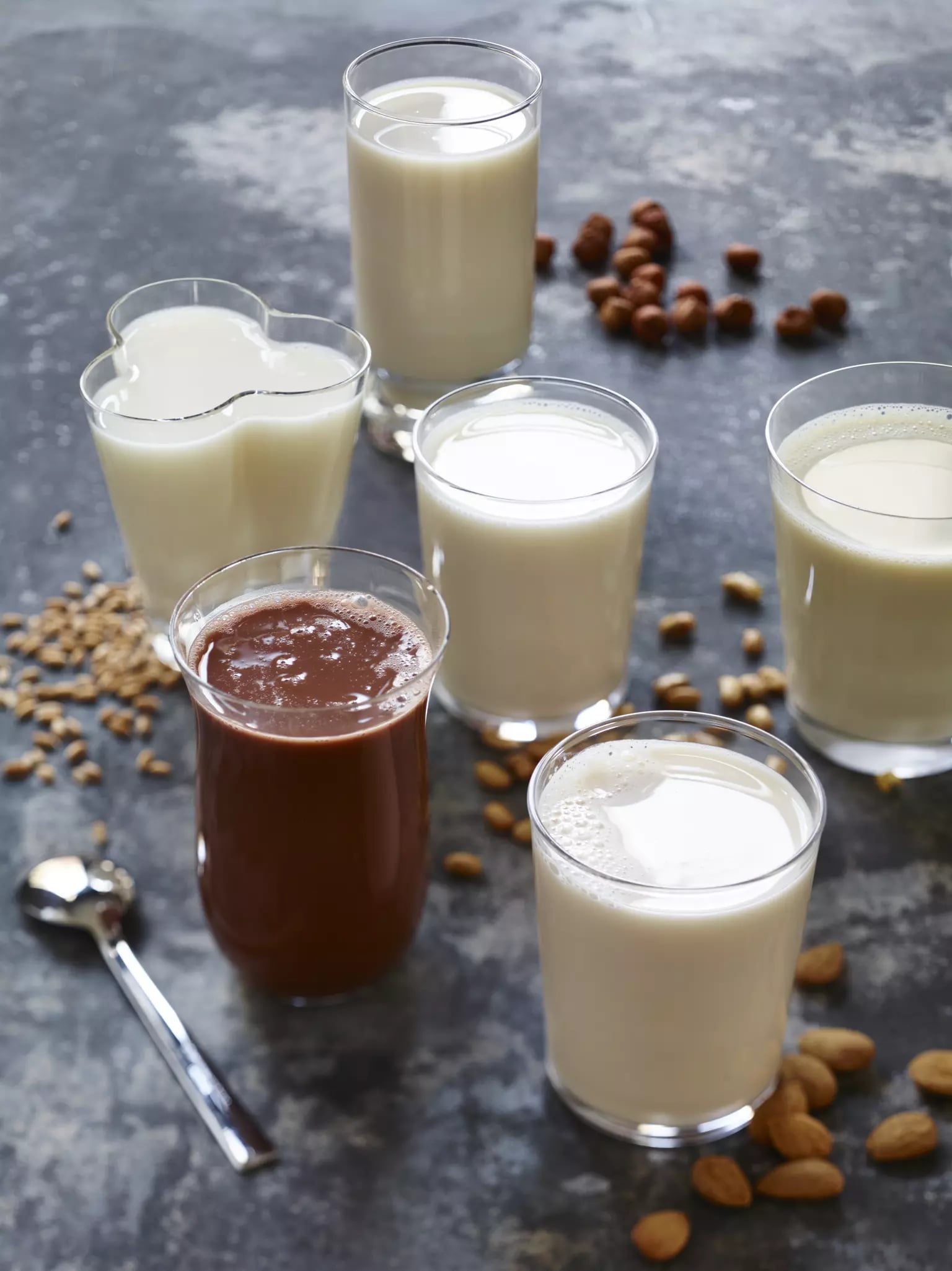 Verschiedene Gläser mit weißen Milchalternativen und ein Glas mit einem braunen pflanzlichen Drink. Neben den Gläsern liegen jeweils die jeweiligen rohen Sorten, wie Mandeln, Haselnüsse, Sojabohnen und Hafer.