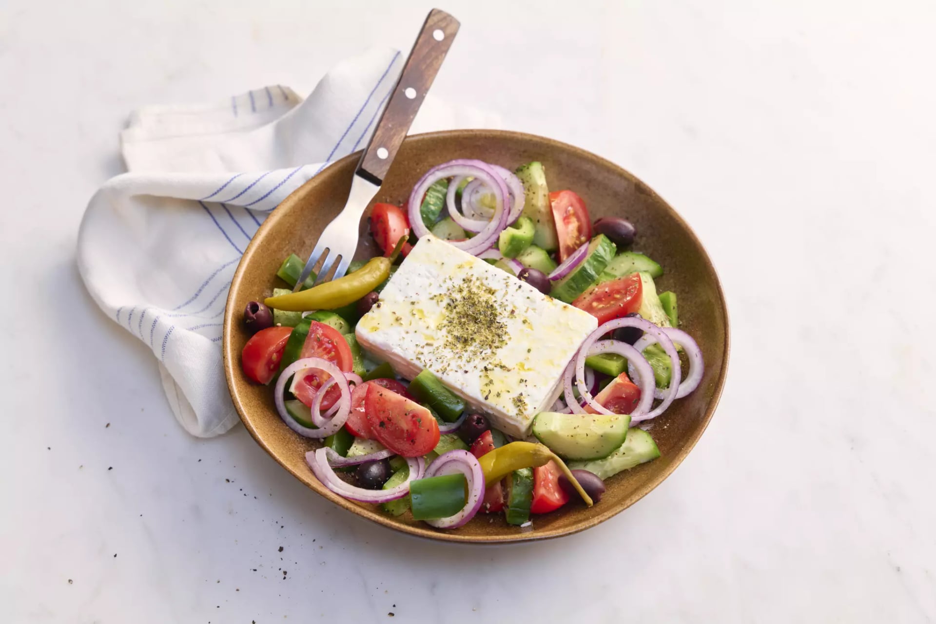 Braune Schale mit griechischem Salat aus Tomaten, Gurken, roten Zwiebelringen, Oliven, Peperoni und einem großen Stück Feta-Käse, daneben ein weiß-blau gestreiftes Küchentuch und eine Gabel mit Holzgriff.