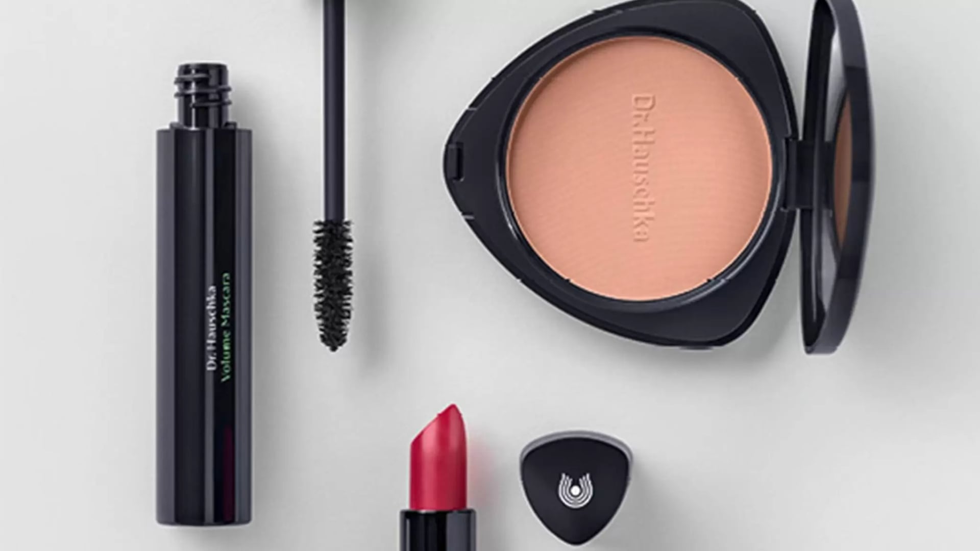 Dr. Hauschka Make-up-Produkte bestehend aus schwarzer Mascara, rotem Lippenstift, beigefarbenem Puder in schwarzer Verpackung und kleinem schwarzem Dreieck mit Dr. Hauscka Logo