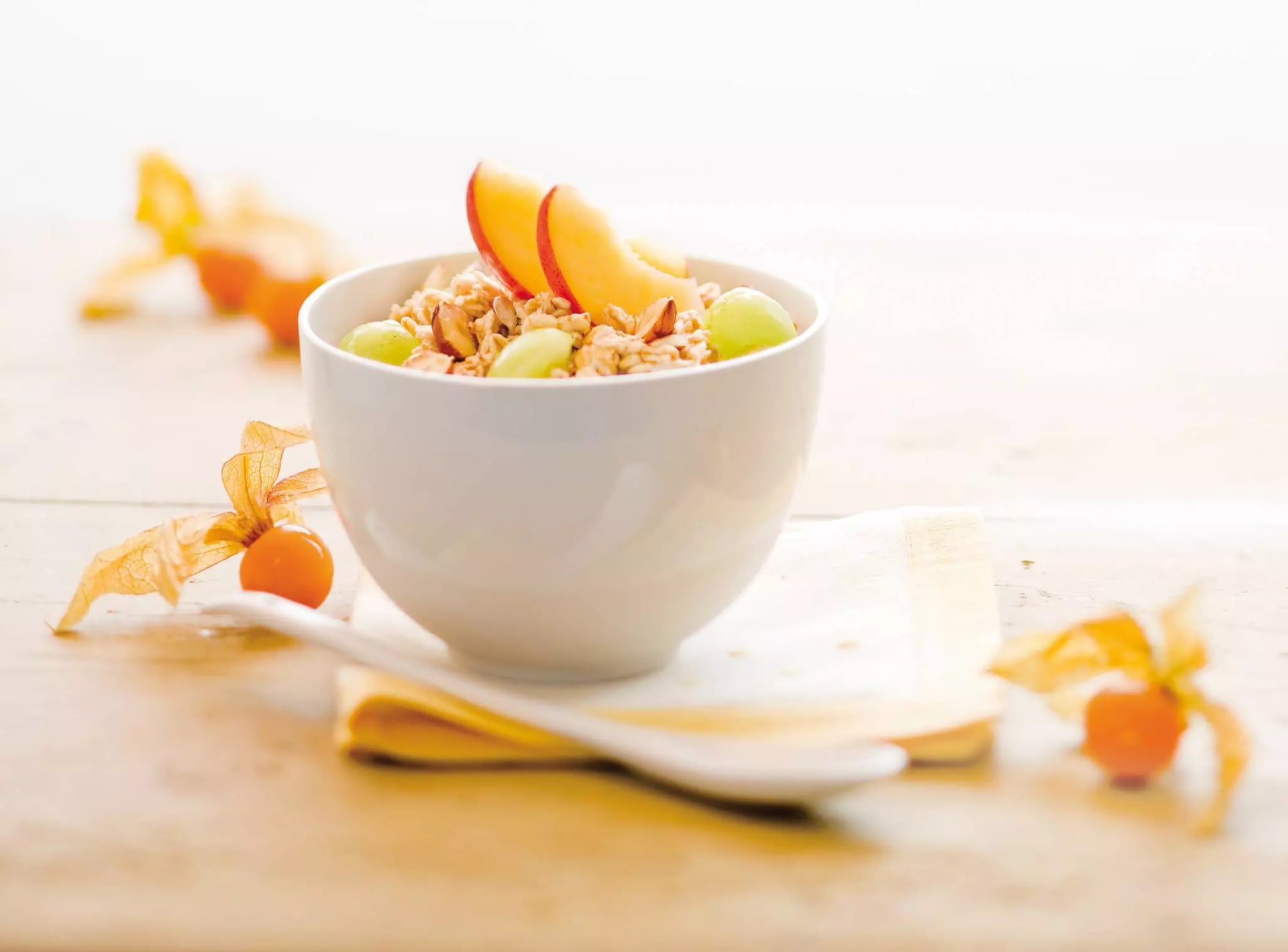 Schale mit Müsli, Trauben, Nüssen und Pfirsichscheiben auf hellem Untergrund mit Physalis-Früchten und weißem Löffel.