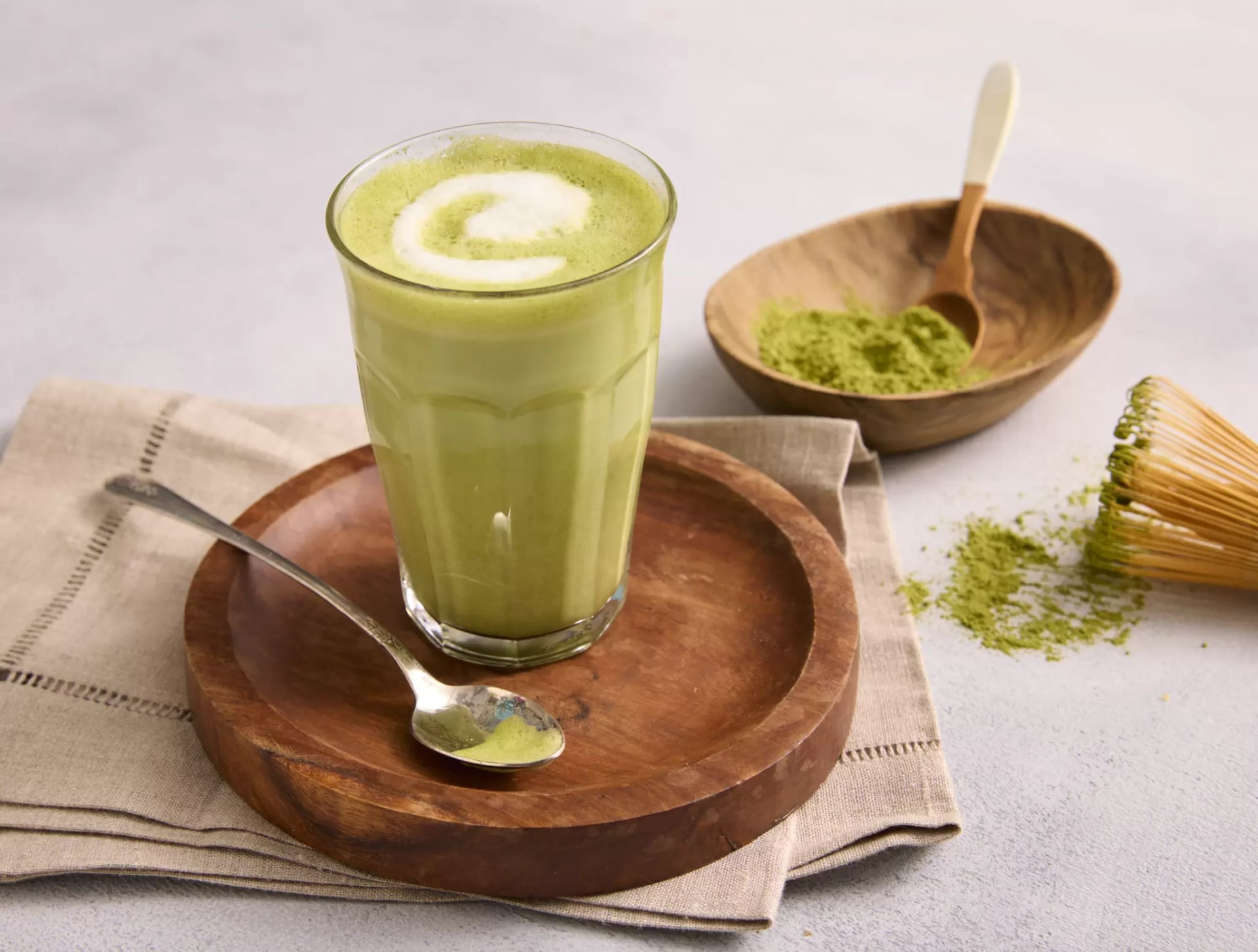 Auf einem hellen Untergrund liegt eine hellbraune Stoffserviette auf dem ein runder dunkler Holzteller steht. Auf dem Holzteller steht ein hohes Glas mit einem grünen Getränk, einem Matcha-Latte. Auf dem Teller liegt ebenfalls ein Teelöffel mit grünem Matchapulver. Im Hintergrund ist eine kleine Holzschüssel mit mehr Matchapulver.
