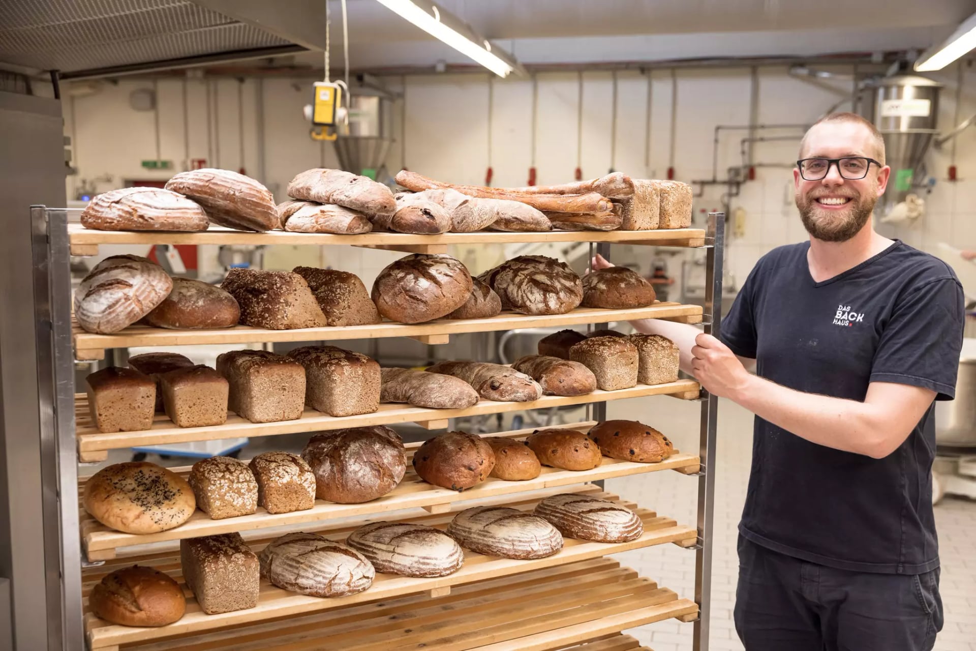 Ein Mann in schwarzem Shirt steht lächelnd neben einem Brot-Rollregal