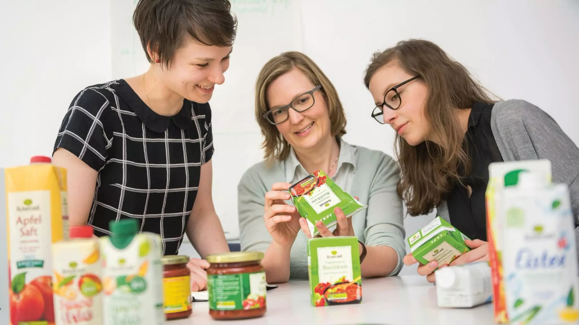 Drei Frauen sitzen an einem Tisch und betrachten gemeinsam Verpackungen von Lebensmitteln in Kartons und Gläsern