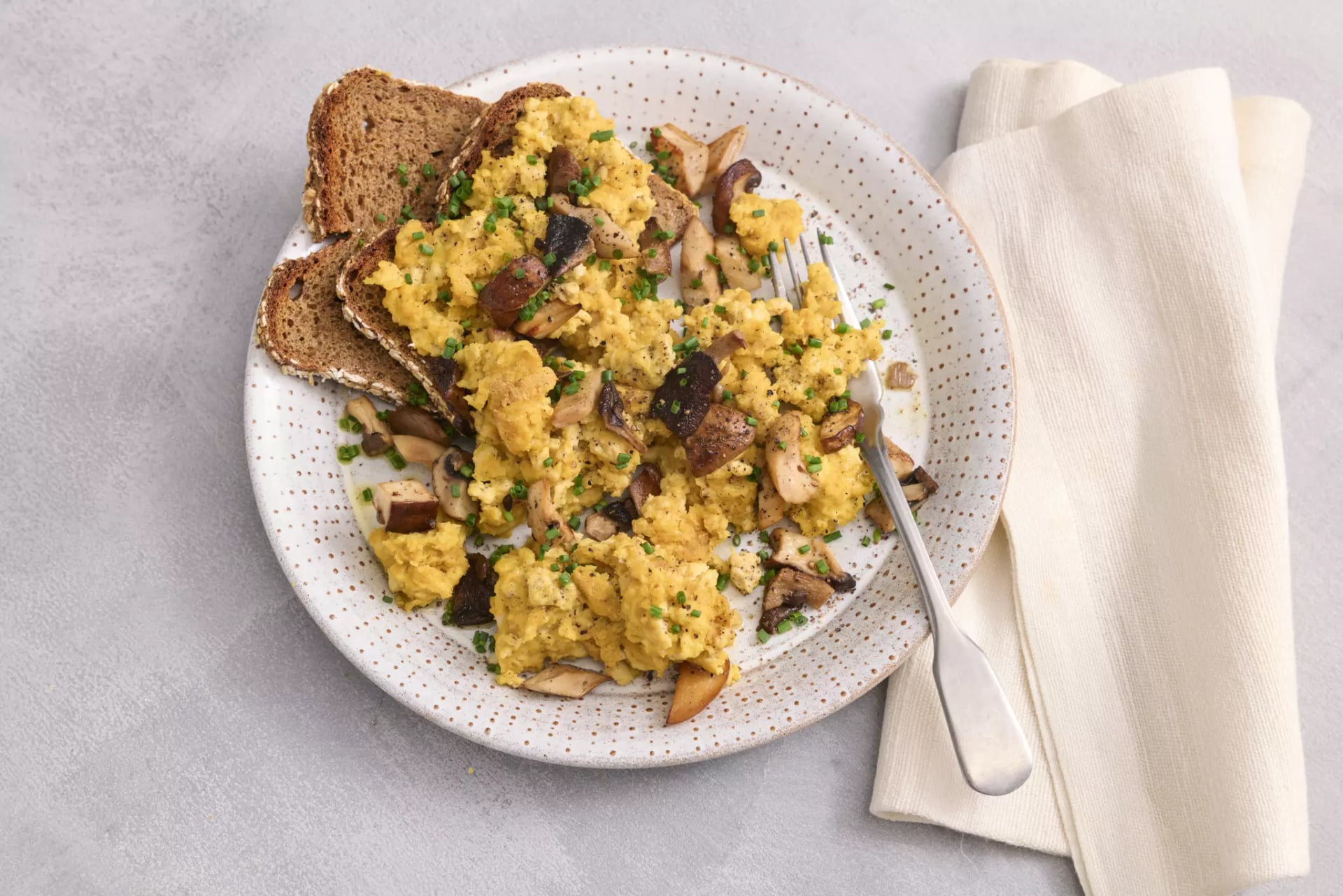 Auf einem weißen Porzellanteller liegt Rührei aus Tofu mit Pilzen und zwei Scheiben dunkles Vollkornbrot. Auf dem Teller liegt noch eine Gabel. Neben dem Teller liegt eine cremefarbene Stoffserviette.