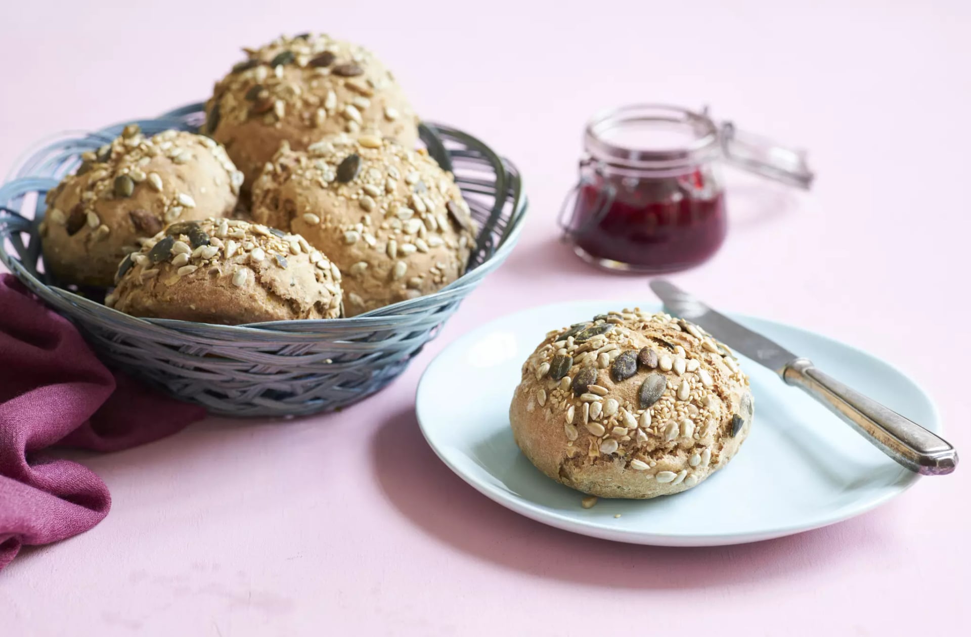 Brötchen mit verschiedenen Kernen in einem Korb und auf einem Teller mit Messer und kleinem Glas mit roter Marmelade auf rosa Untergrund.